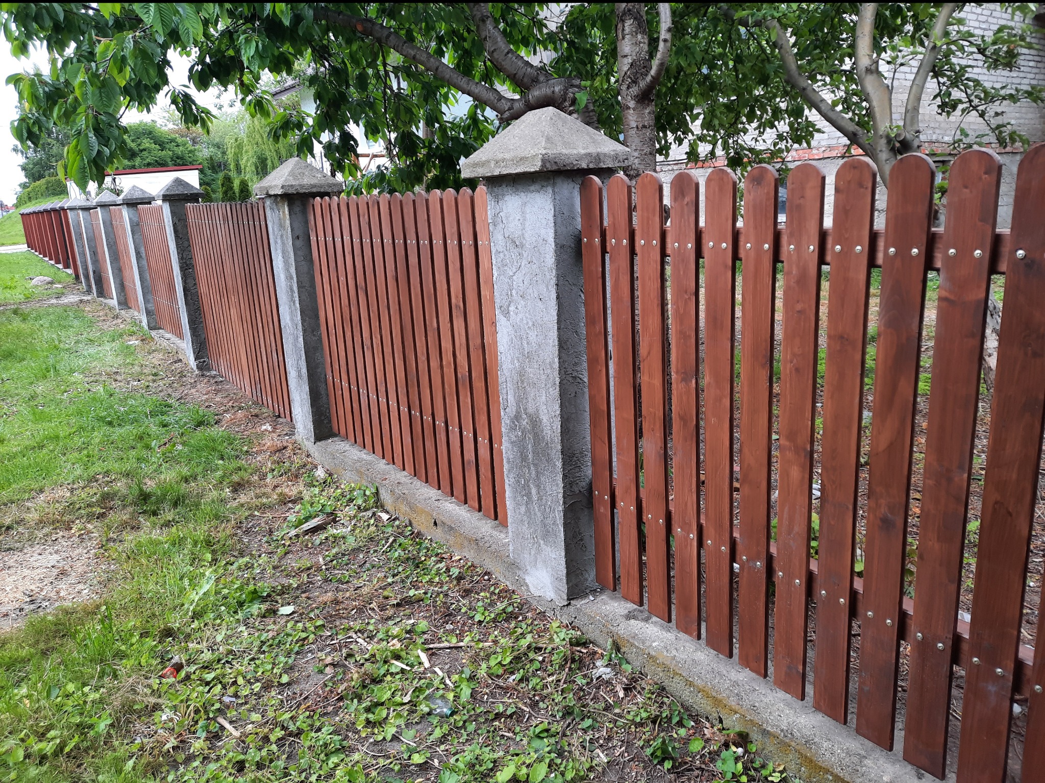 Długi, niski płot z brązowych desek i betonowych słupków z piramidkami, biegnący wzdłuż trawnika. Widok z boku, w tle zieleń drzew i fragment budynku.