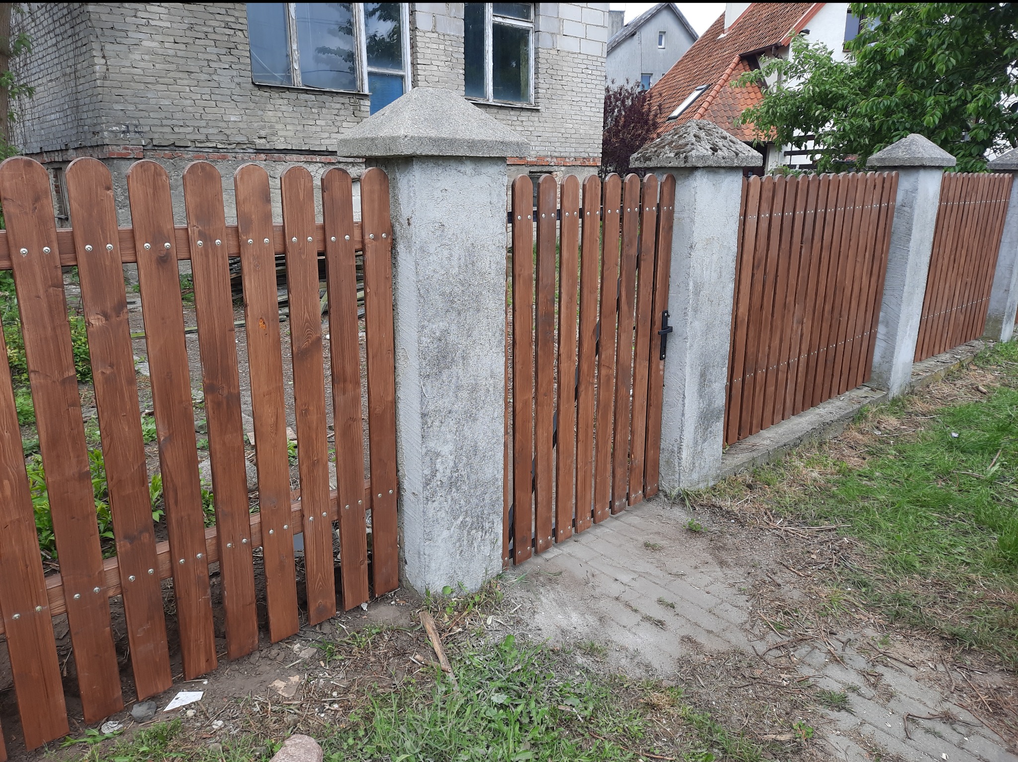 Drewniane ogrodzenie z furtką, brązowe deski pionowe, szare betonowe słupki z daszkami, tło: budynek z szarej cegły i dom z czerwonym dachem.