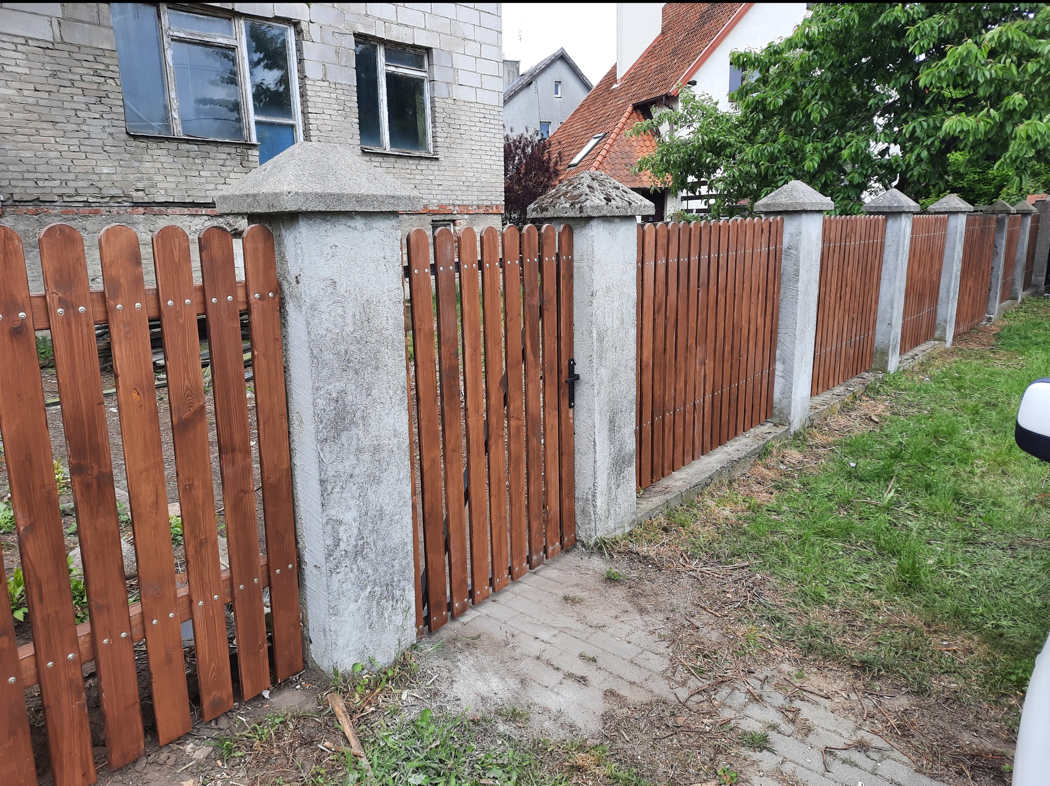 Nowe ogrodzenie palisadowe z drewnianymi sztachetami w ciepłym odcieniu brązu, zamontowane na betonowych słupkach z dekoracyjnymi daszkami, otacza posesję z budynkami w tle.