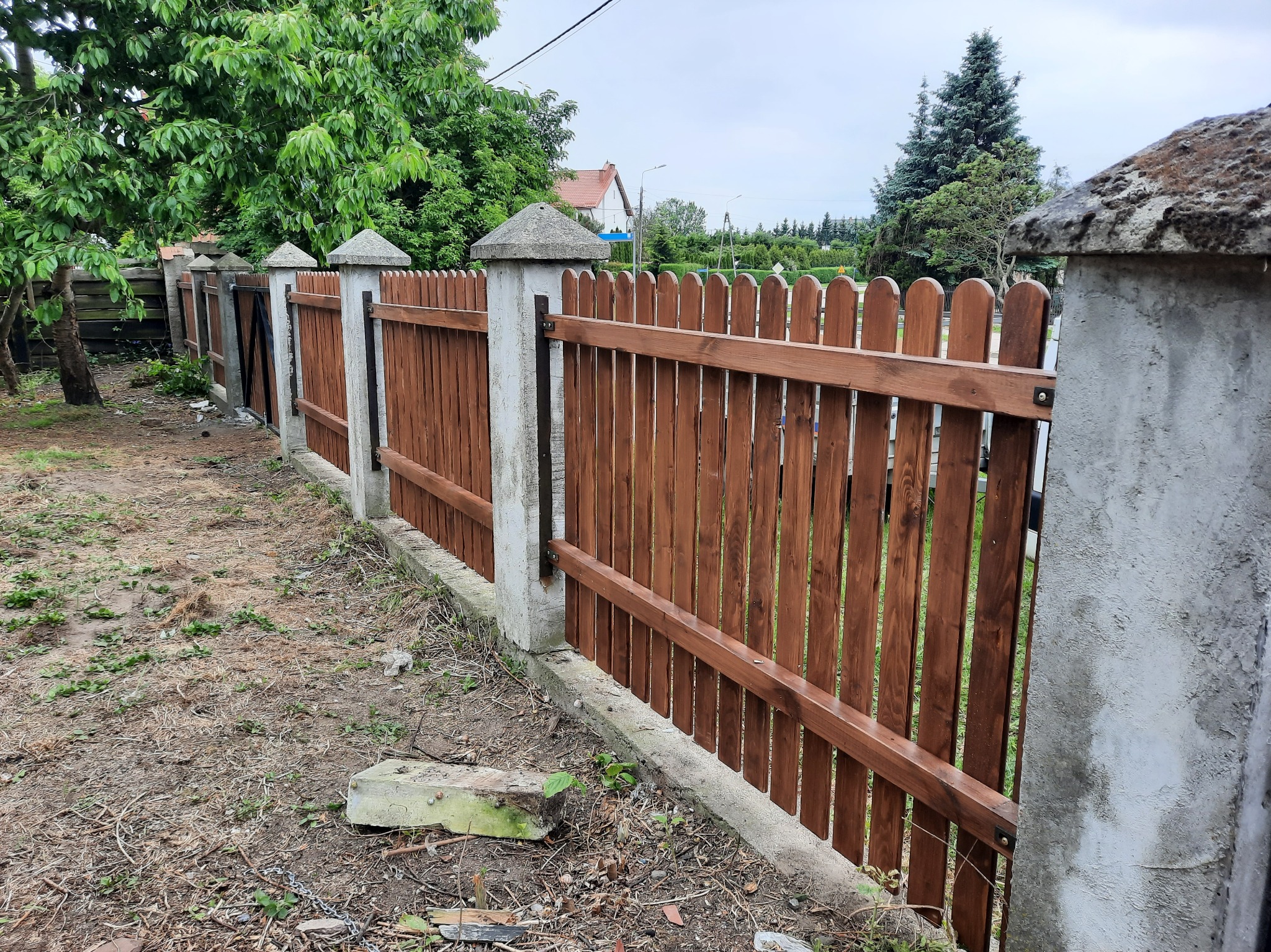 Drewniane ogrodzenie z betonowymi słupkami i daszkami, pomalowane na brązowo, z widocznymi elementami montażowymi, otoczone zielenią. Widok z perspektywy.