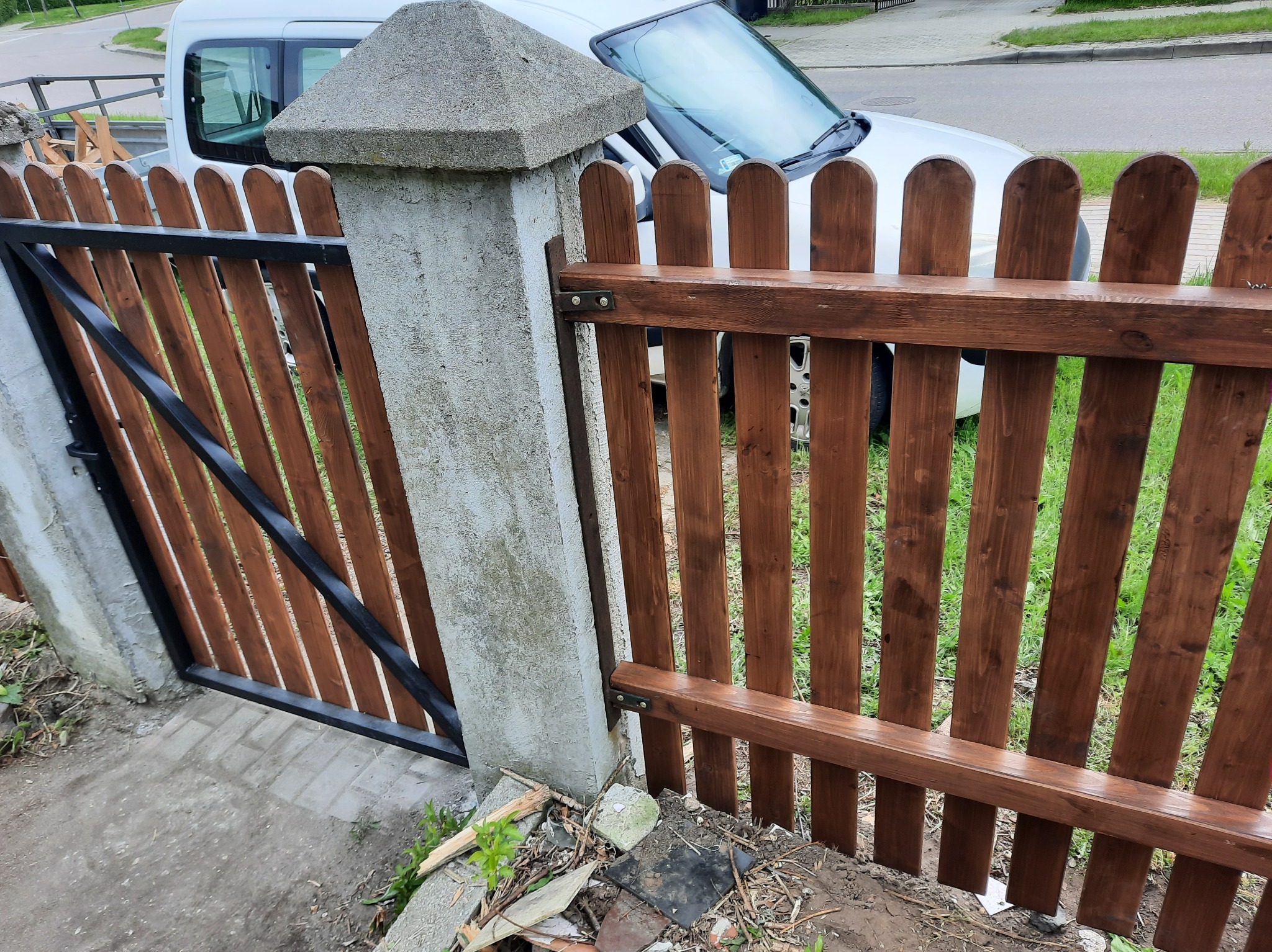 Świeżo pomalowane drewniane ogrodzenie z bramką, widoczne słupki betonowe i fragment samochodu w tle. Ujęcie z niskiej perspektywy.