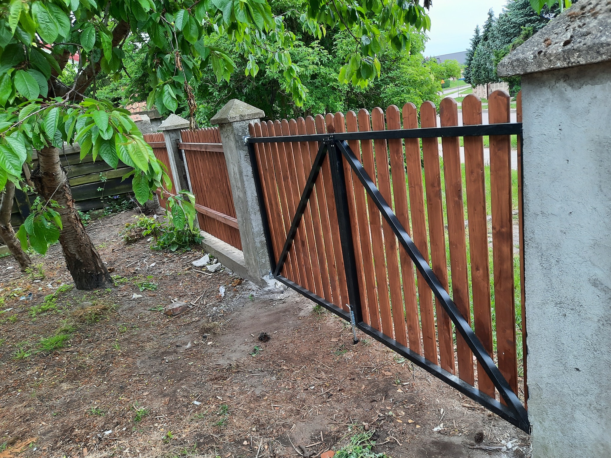 Otwarta furtka z drewnianymi sztachetami i metalową ramą, osadzona w betonowych słupkach. Widok z zewnątrz posesji, zieleń w tle.