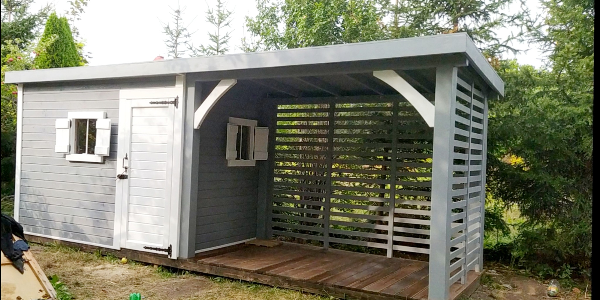 Szara altana z drewnianą podłogą i ażurowymi ścianami, zamykana drzwiami i oknem z okiennicami, na tle zieleni ogrodu.