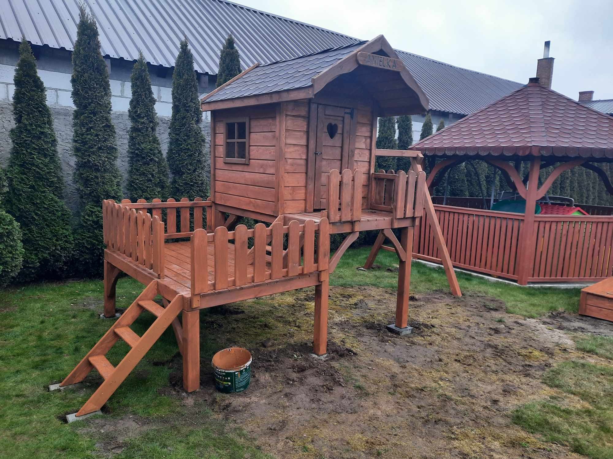 Drewniany domek dla dzieci na podwyższeniu z balustradą, schodami i małą werandą. Obok widoczna altana i wysokie tuje.