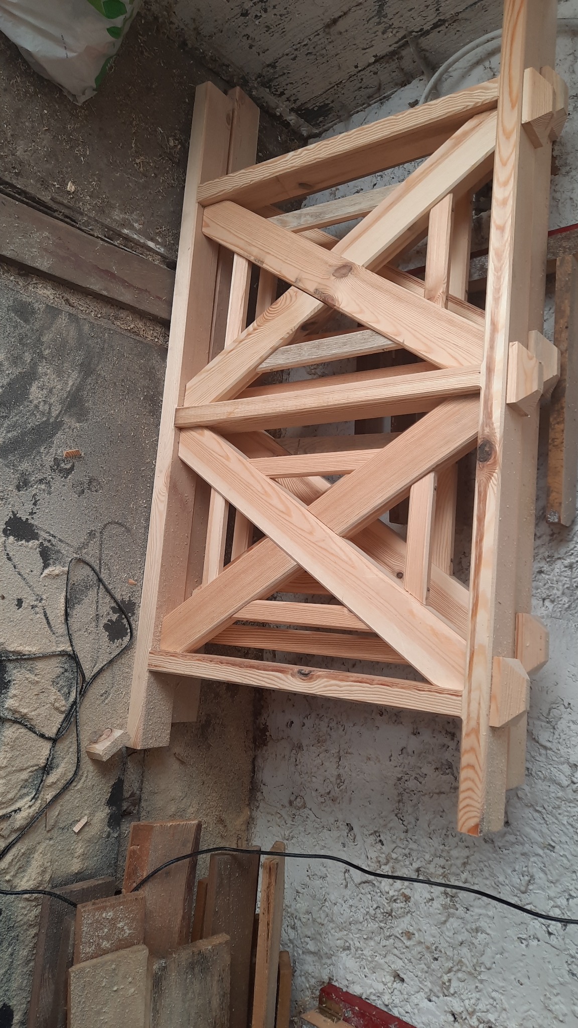 Surowa, drewniana konstrukcja z krzyżującymi się belkami, prawdopodobnie element balustrady. Widoczny fragment ściany i podłogi z resztkami obróbki drewna.