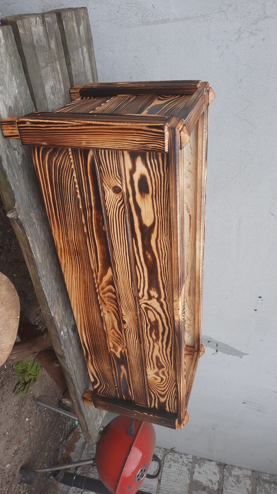 Ręcznie wykonana drewniana skrzynia z efektem opalania, widoczne słoje drewna i dekoracyjne wykończenia, leżąca na boku obok czerwonego grilla.