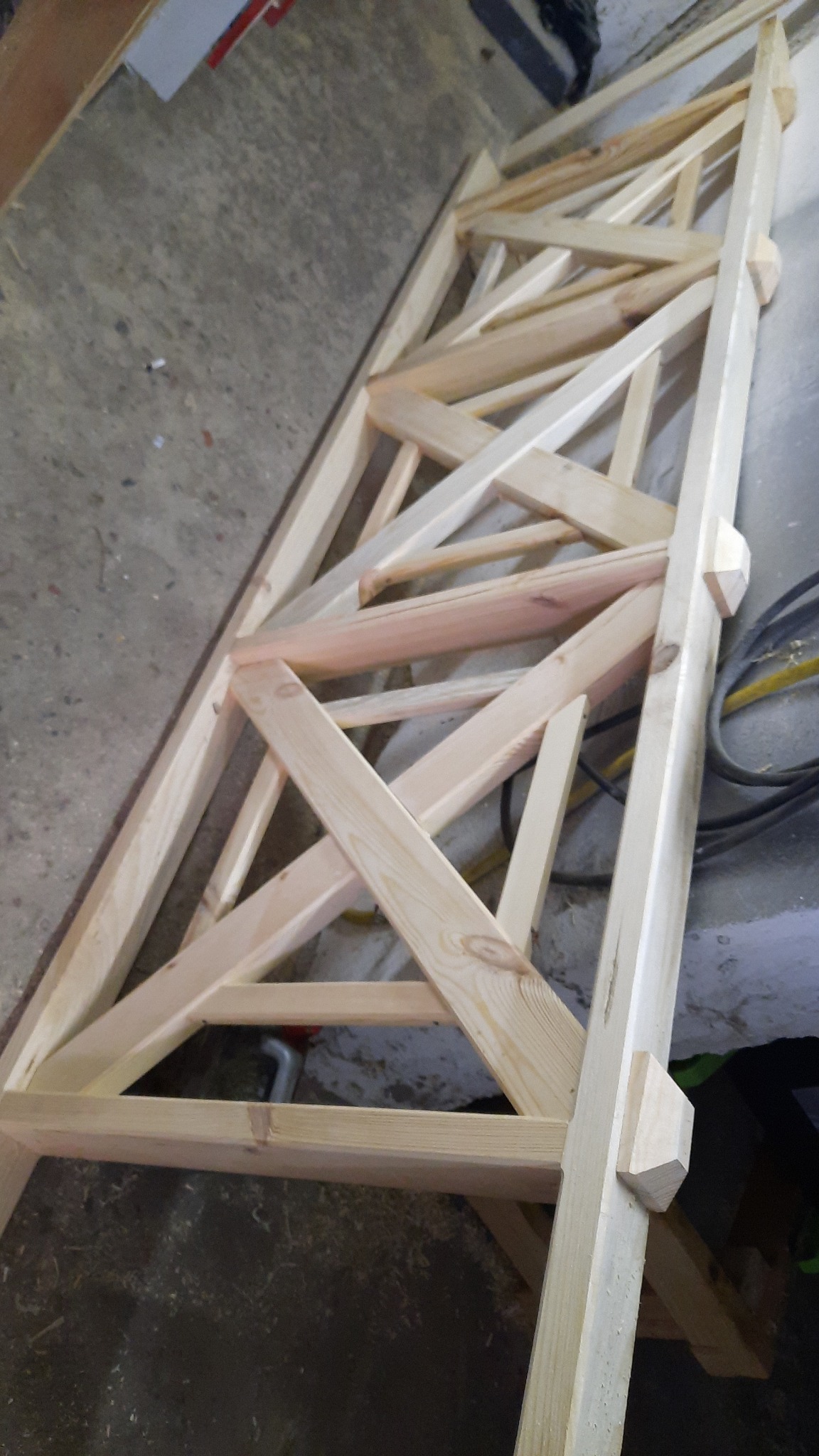 Surowa drewniana konstrukcja z belek, złożona w kratownicę, leżąca na podłodze. Widoczne precyzyjne łączenia elementów i naturalny odcień drewna.