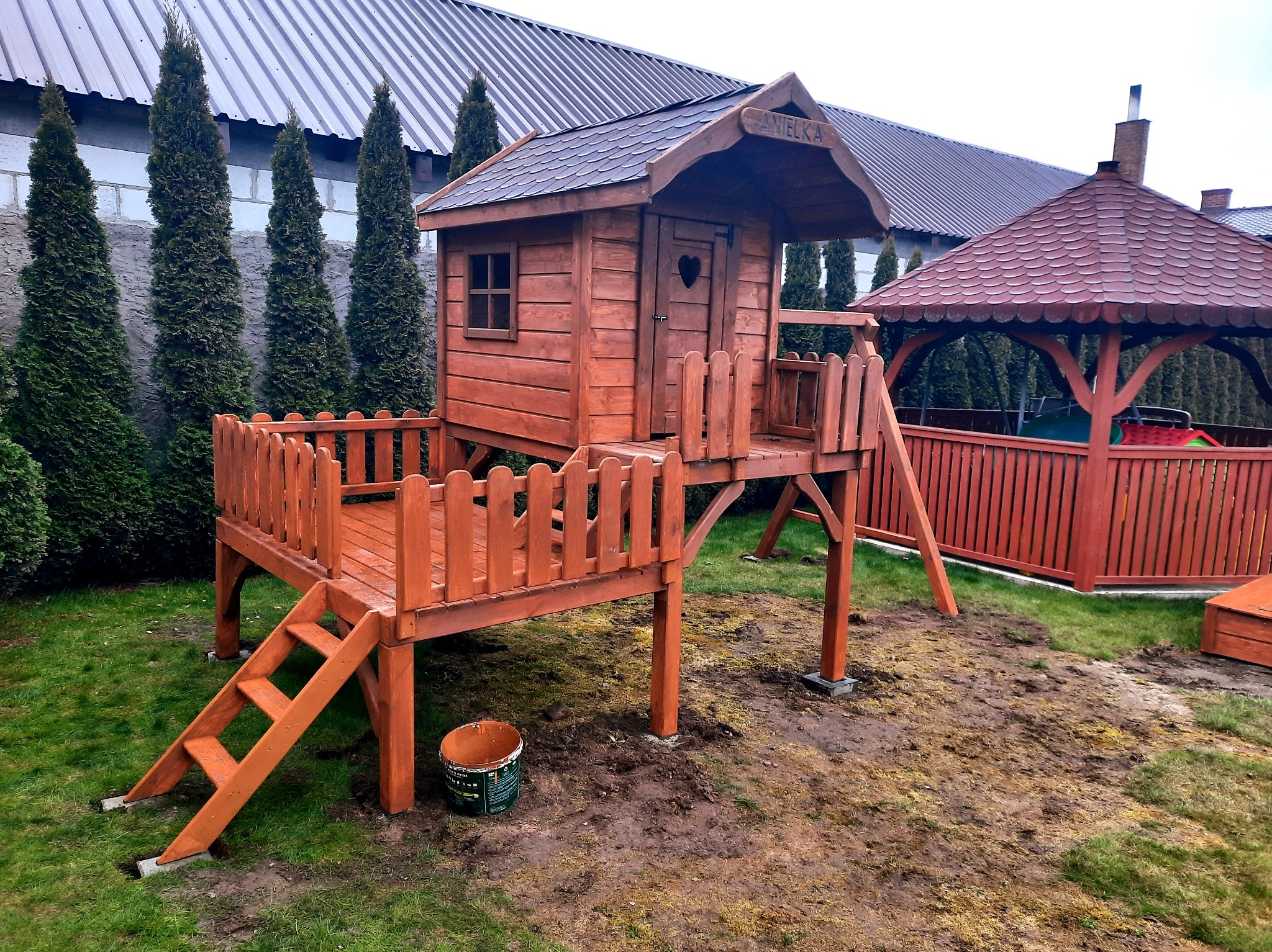 Drewniany plac zabaw dla dzieci z domkiem na podeście i drabinką, pomalowany na brązowo, z widoczną altaną w tle. Na domku widnieje napis 'Anielka'.