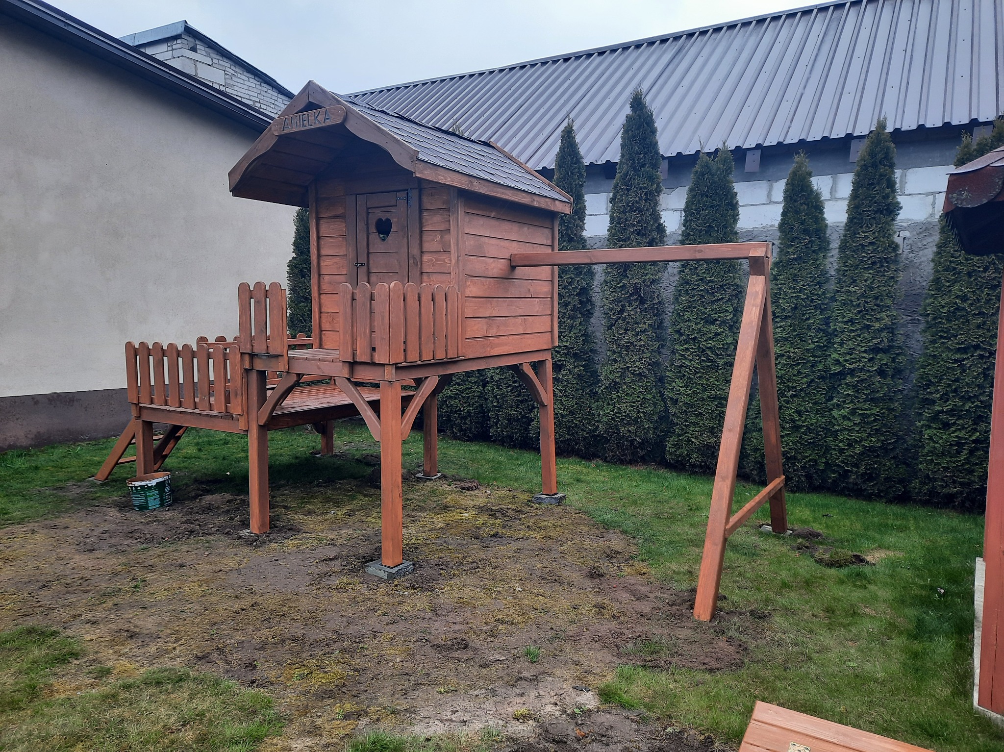 Drewniany domek dla dzieci z huśtawką, ustawiony w ogrodzie. Domek z dachem krytym gontem, na podwyższeniu, z małym balkonem i schodkami. Widoczne drzewa w tle.