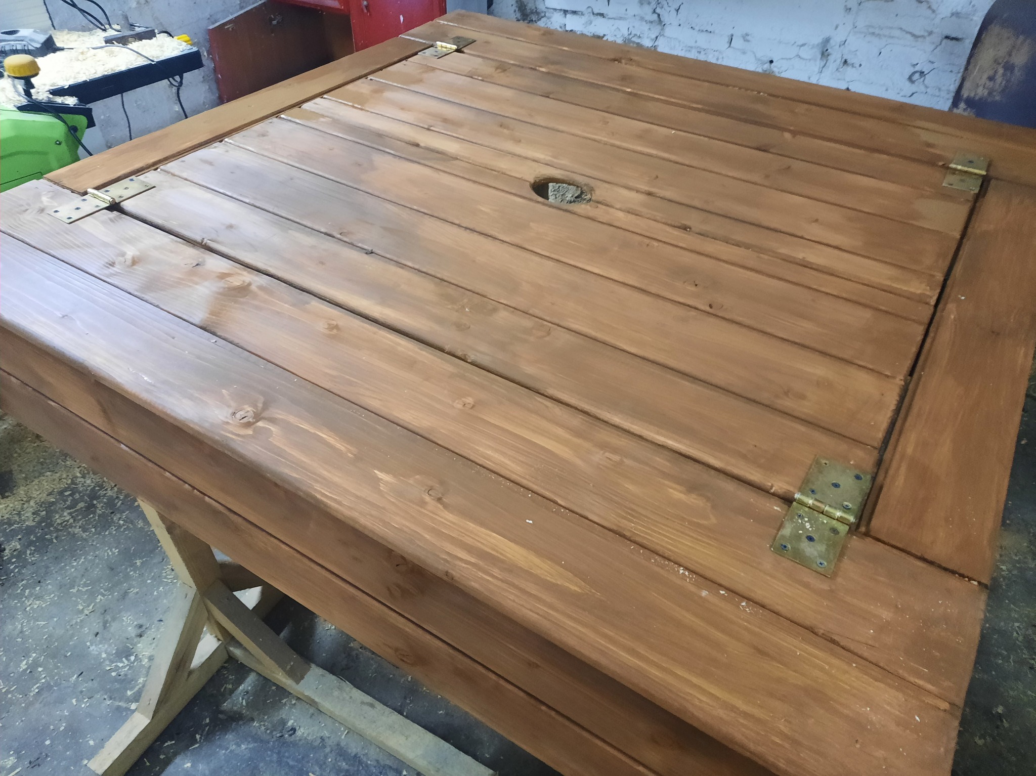 Drewniany stół z otworem na parasol, wykonany z desek w ciepłym, brązowym odcieniu, wsparty na drewnianej konstrukcji. Widoczne zawiasy i ślady obróbki.