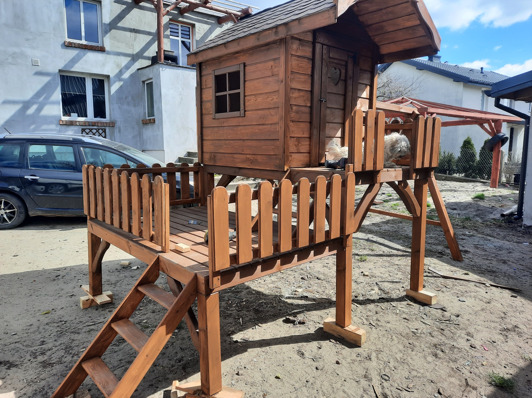 Drewniany domek dla dzieci na podwyższeniu z balustradą i schodkami, umieszczony na trawniku przed budynkiem mieszkalnym. Widoczny pies w domku.