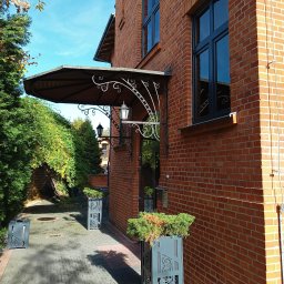 KANCELARIA ADWOKACKA ADWOKAT NORBERT PODDĘBSKI - Eleganckie wejście do budynku w Łodzi, z ceglaną elewacją, ozdobnym daszkiem nad drzwiami, metalowymi donicami z zielenią i brukowanym chodnikiem.