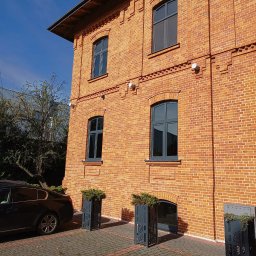 KANCELARIA ADWOKACKA ADWOKAT NORBERT PODDĘBSKI - Odrestaurowana kamienica z czerwonej cegły z szarymi oknami i dekoracyjnymi donicami przy wejściu, widok z dołu w słoneczny dzień.