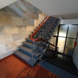 KANCELARIA ADWOKACKA ADWOKAT NORBERT PODDĘBSKI - Klatka schodowa z drewnianą poręczą i ozdobnymi metalowymi balustradami, ściany wyłożone kamieniem, widoczne drzwi z szybą i gaśnica.