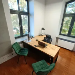 Nowoczesne biuro z dużym drewnianym biurkiem, laptopem, monitorem, zielonymi krzesłami i widokiem z okna na drzewa.