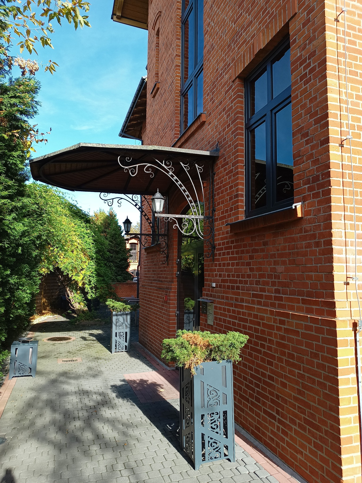 Eleganckie wejście do budynku w Łodzi, z ceglaną elewacją, ozdobnym daszkiem nad drzwiami, metalowymi donicami z zielenią i brukowanym chodnikiem.