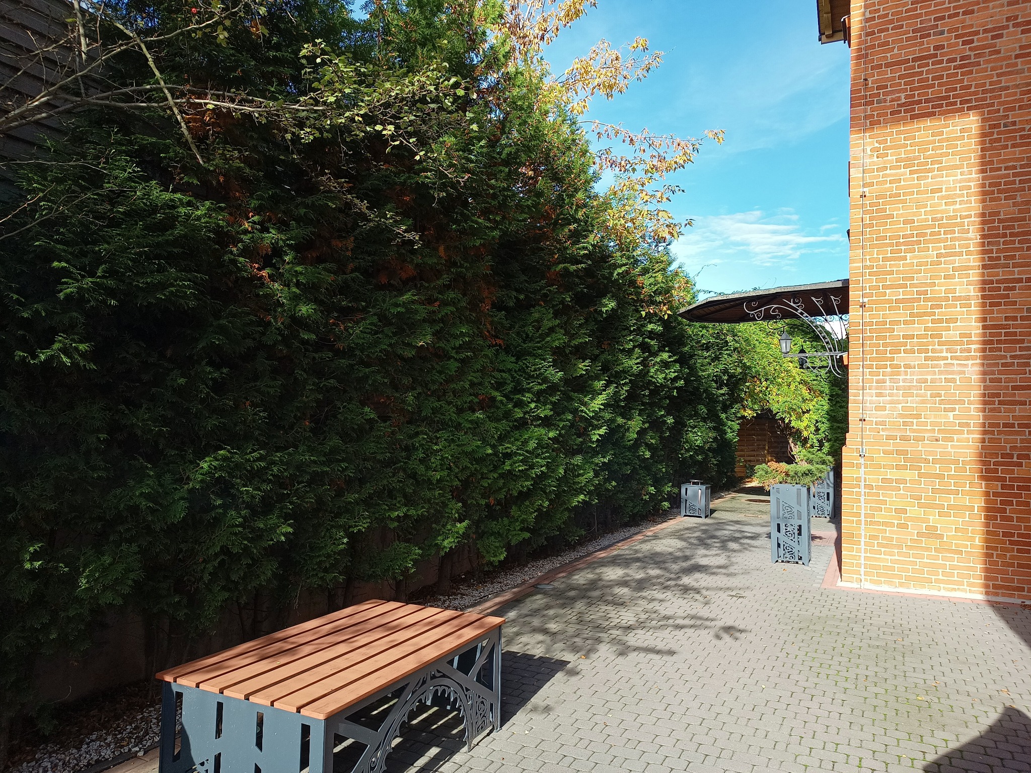 Ustawiona na brukowanym podwórku ławka z drewnianym siedziskiem i metalową konstrukcją, w tle wysokie tuje i ceglany budynek w słoneczny dzień.