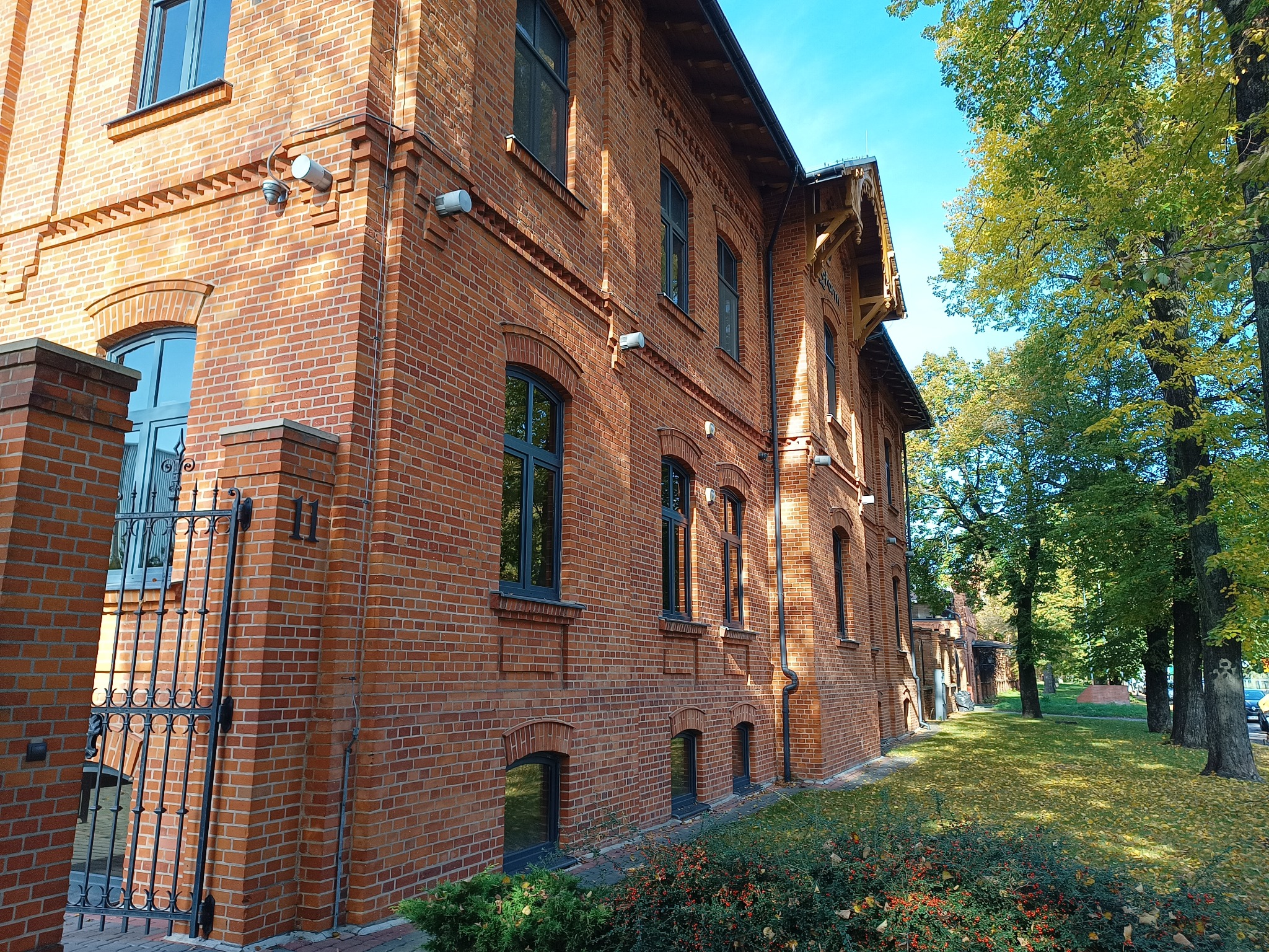 Ceglany budynek z ciemnymi ramami okiennymi, widoczny numer '11' przy wejściu, metalowa brama, otoczenie zieleni i drzew w jesiennej aurze.