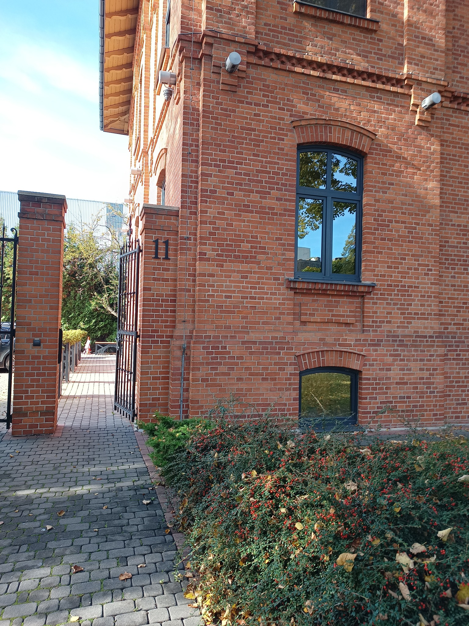 Ceglany budynek z numerem 11, widok z zewnątrz, brama wjazdowa i zadbana zieleń przy budynku.