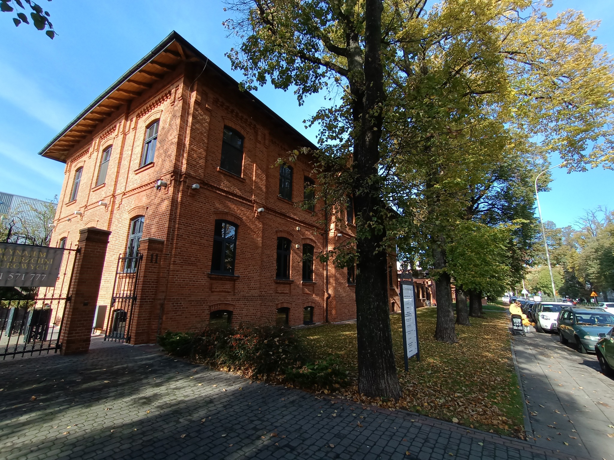 Ceglany budynek biurowy z czarnymi oknami, otoczony drzewami i chodnikiem z kostki, w tle błękitne niebo.