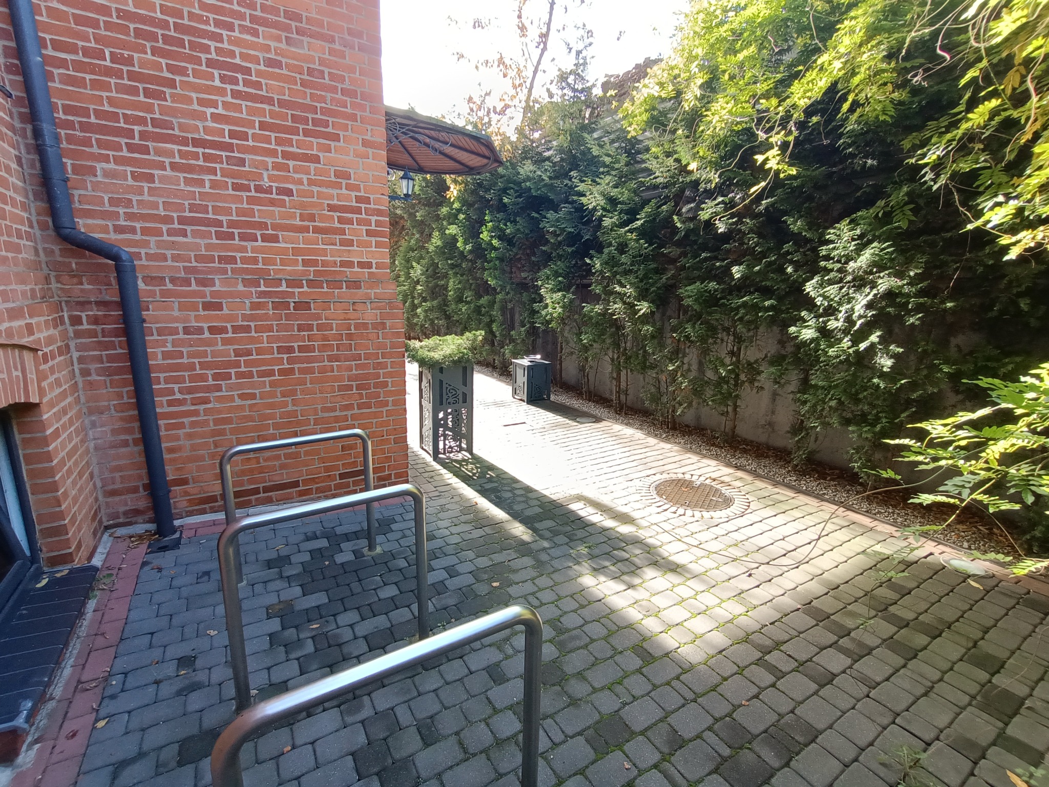 Ujęcie z poziomu gruntu na ścieżkę z kostki brukowej, obok budynku z cegły i metalowych stojaków na rowery, prowadzącą w kierunku zieleni i ozdobnych donic.