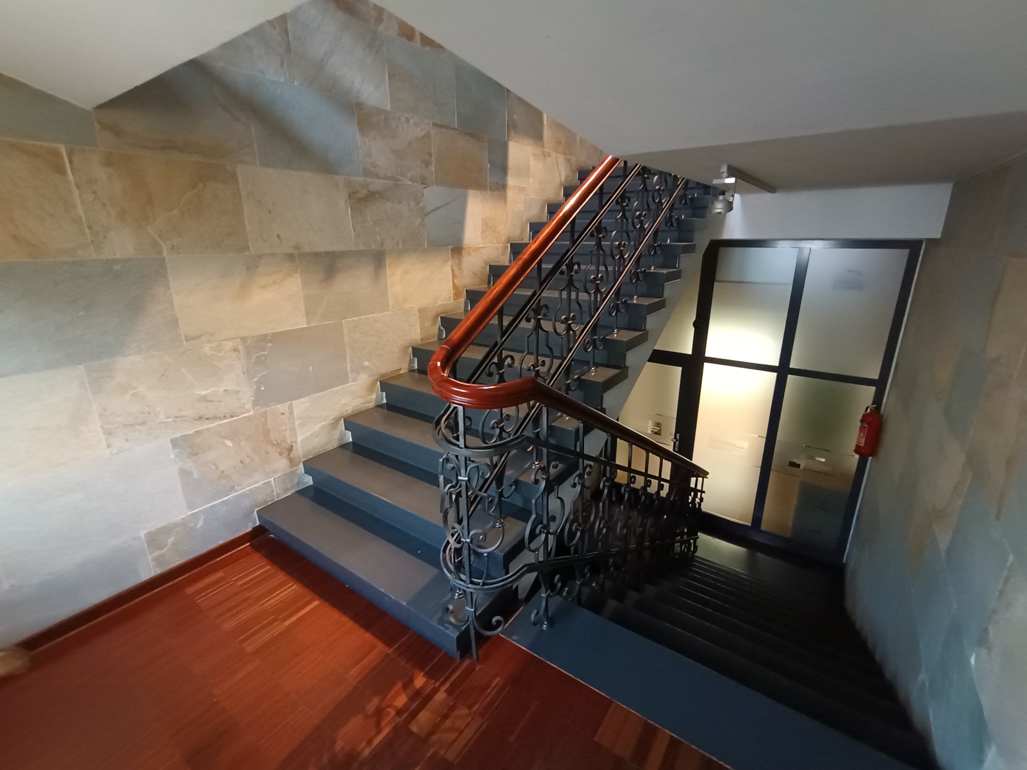 Klatka schodowa z drewnianą poręczą i ozdobnymi metalowymi balustradami, ściany wyłożone kamieniem, widoczne drzwi z szybą i gaśnica.