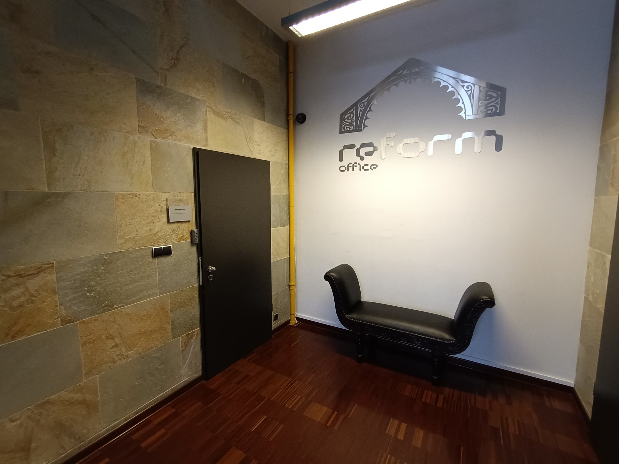 Wnętrze biura z kamienną ścianą, ciemnymi drzwiami, drewnianą podłogą, czarną ławką i logo firmy na białej ścianie.