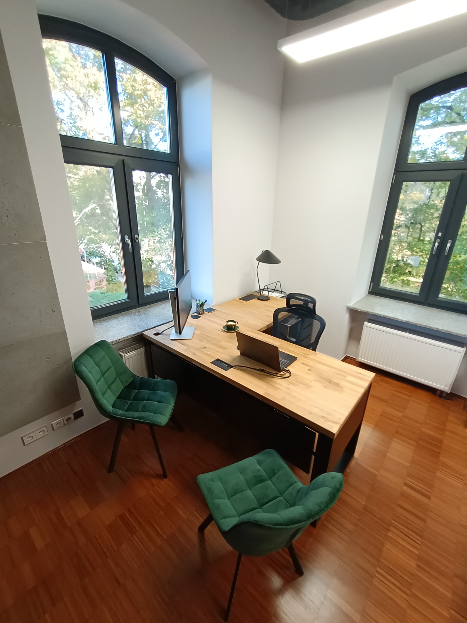 Nowoczesne biuro z dużym drewnianym biurkiem, laptopem, monitorem, zielonymi krzesłami i widokiem z okna na drzewa.