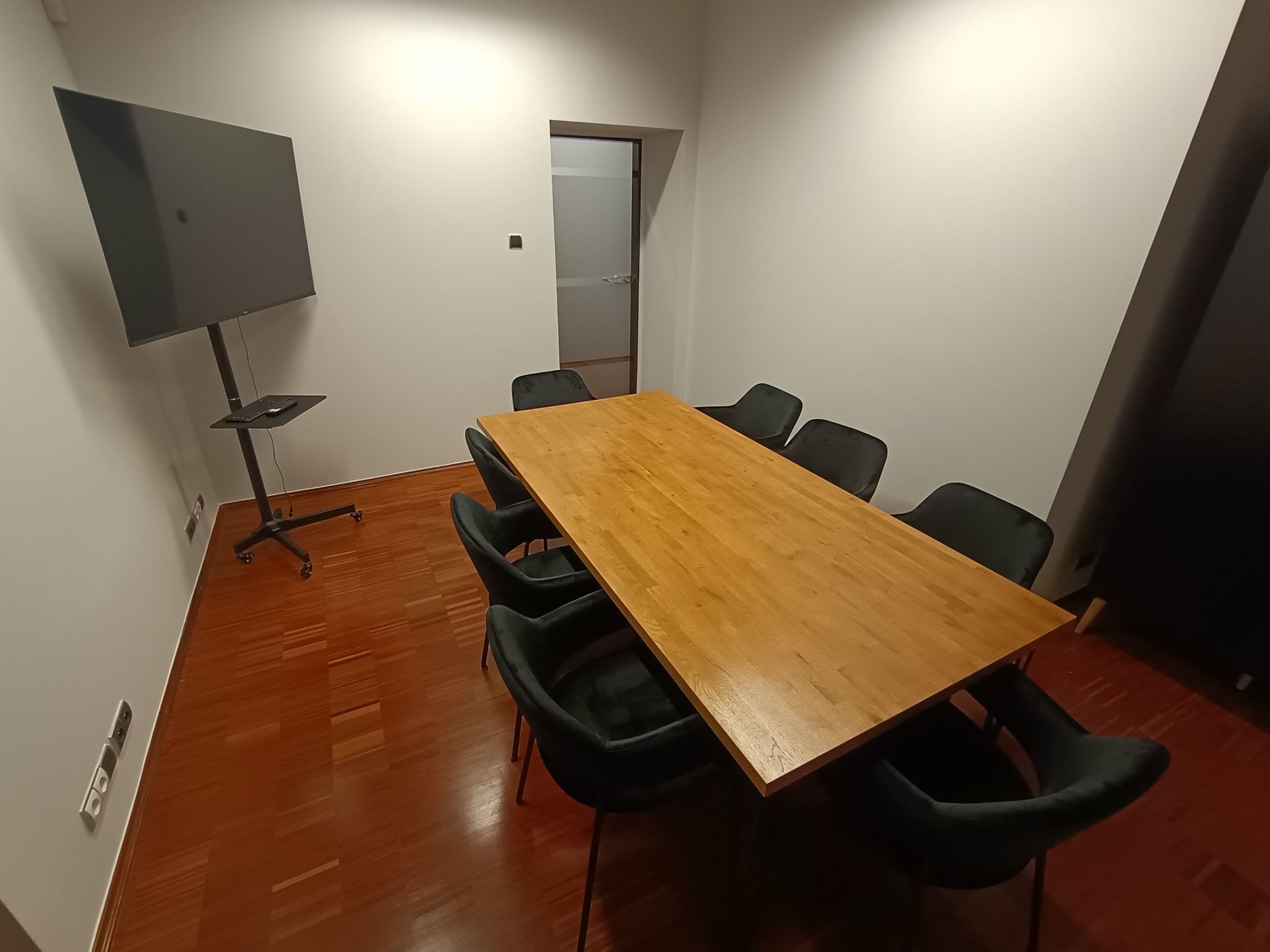 Sala konferencyjna z drewnianym stołem, czarnymi krzesłami i dużym ekranem na statywie. Widoczne gniazdka elektryczne w ścianie i drzwi w tle.