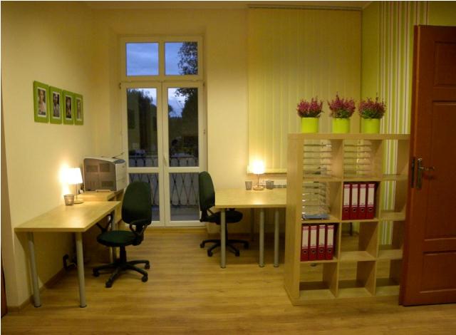 Jasne wnętrze biura z dwoma stanowiskami pracy, biurkami, krzesłami obrotowymi, regałem z segregatorami i roślinami w doniczkach, z widokiem na balkon.