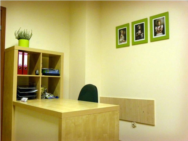 Jasne biuro z biurkiem, regałem z segregatorami i trzema portretami w zielonych ramkach na ścianie, składany blat przymocowany do ściany.