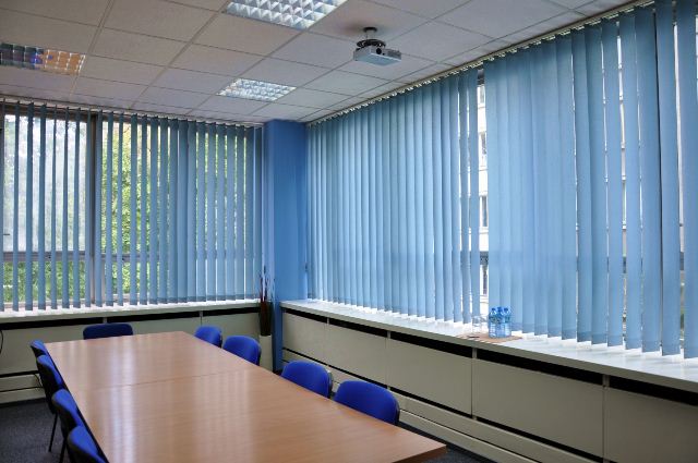 Jasna sala konferencyjna z długim stołem, niebieskimi krzesłami, pionowymi żaluzjami w oknach i projektorem na suficie.