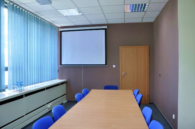 Sala konferencyjna z projektorem, długim stołem z niebieskimi krzesłami, zasłonięte okno z pionowymi żaluzjami.