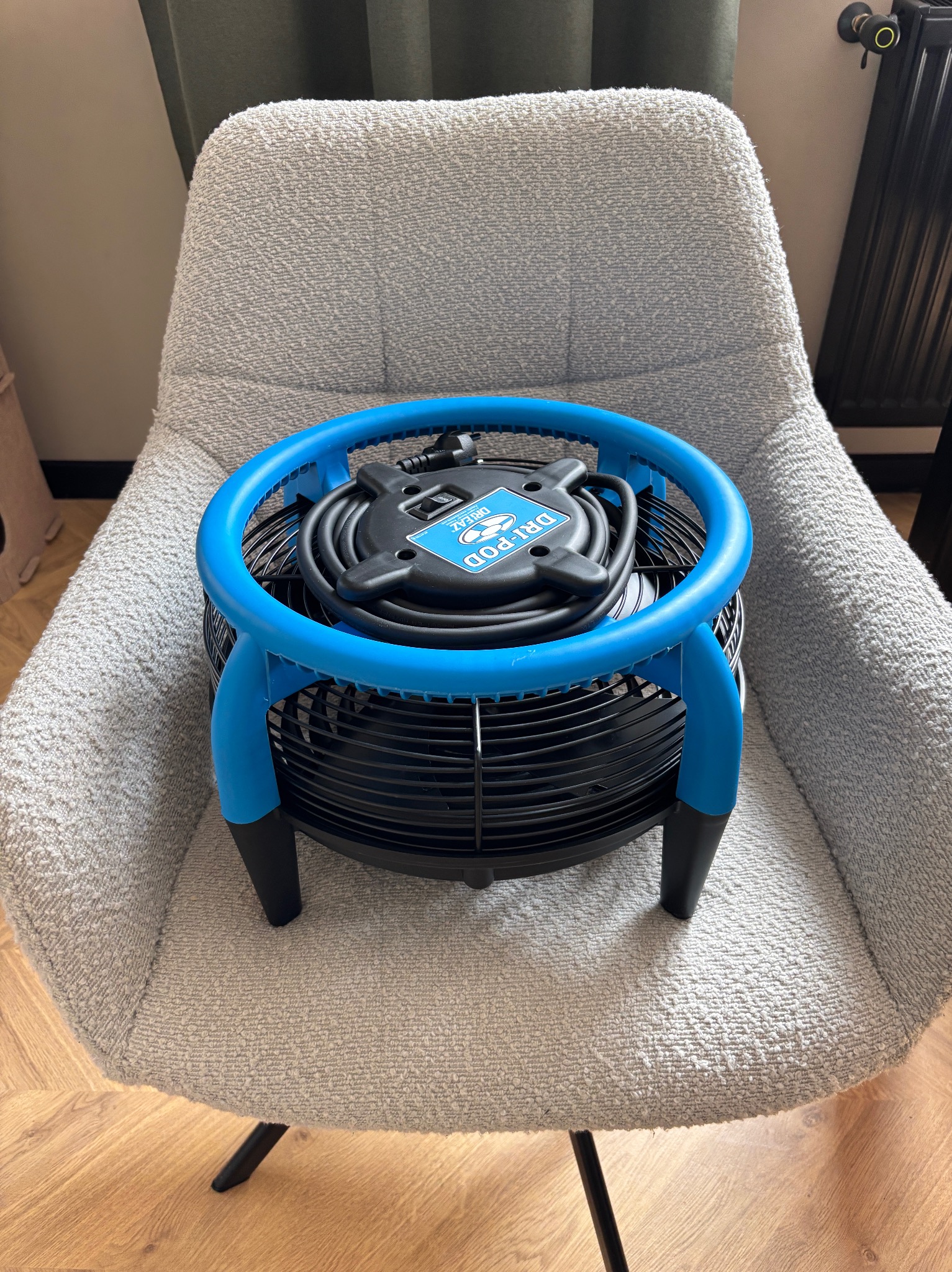 Dri-Pod wentylator osuszający, czarny z niebieską obudową, leży na szarym fotelu z tkaniny boucle. Widoczny zwinięty kabel. Na podłodze panele.