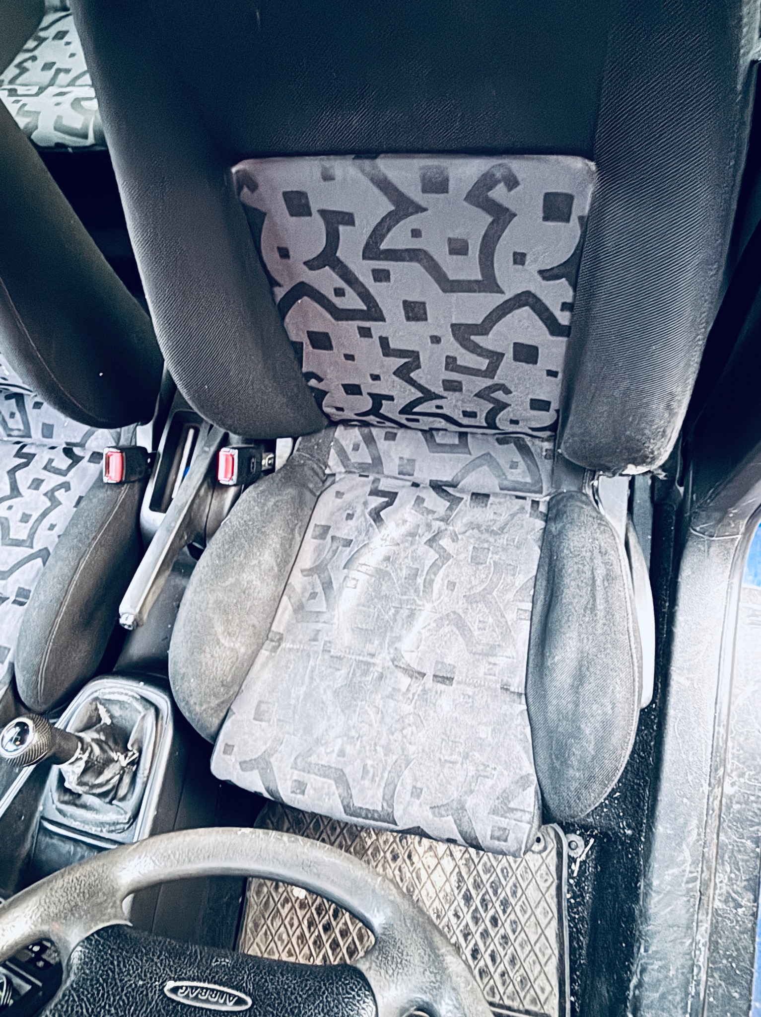 Brudne siedzenie samochodu z wzorem geometrycznym, widoczne elementy wnętrza pojazdu, kierownica, dźwignia zmiany biegów. Fotel wymaga czyszczenia.
