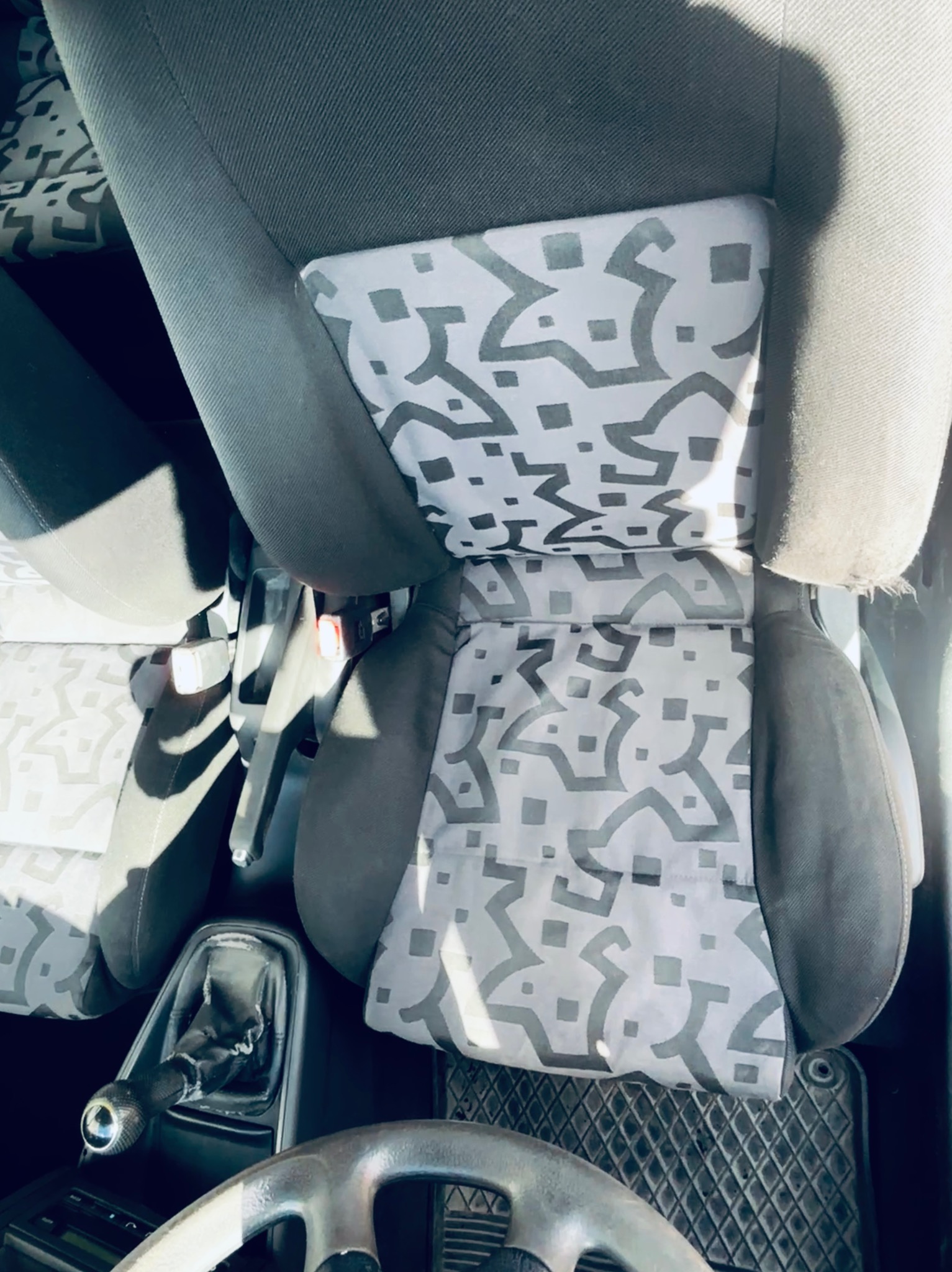Wnętrze samochodu: fotel kierowcy z szaro-białą tapicerką w geometryczne wzory, widoczna kierownica i dźwignia zmiany biegów. Ujęcie z góry.