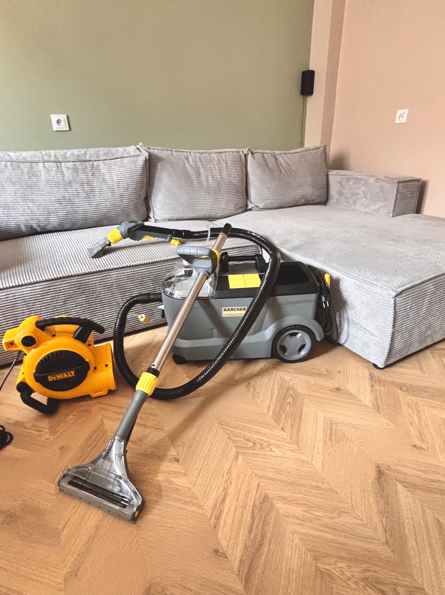 Sprzęt do czyszczenia tapicerki Karcher i DeWalt obok narożnika z szarej tkaniny w paski, na parkiecie ułożonym we wzór jodełki.