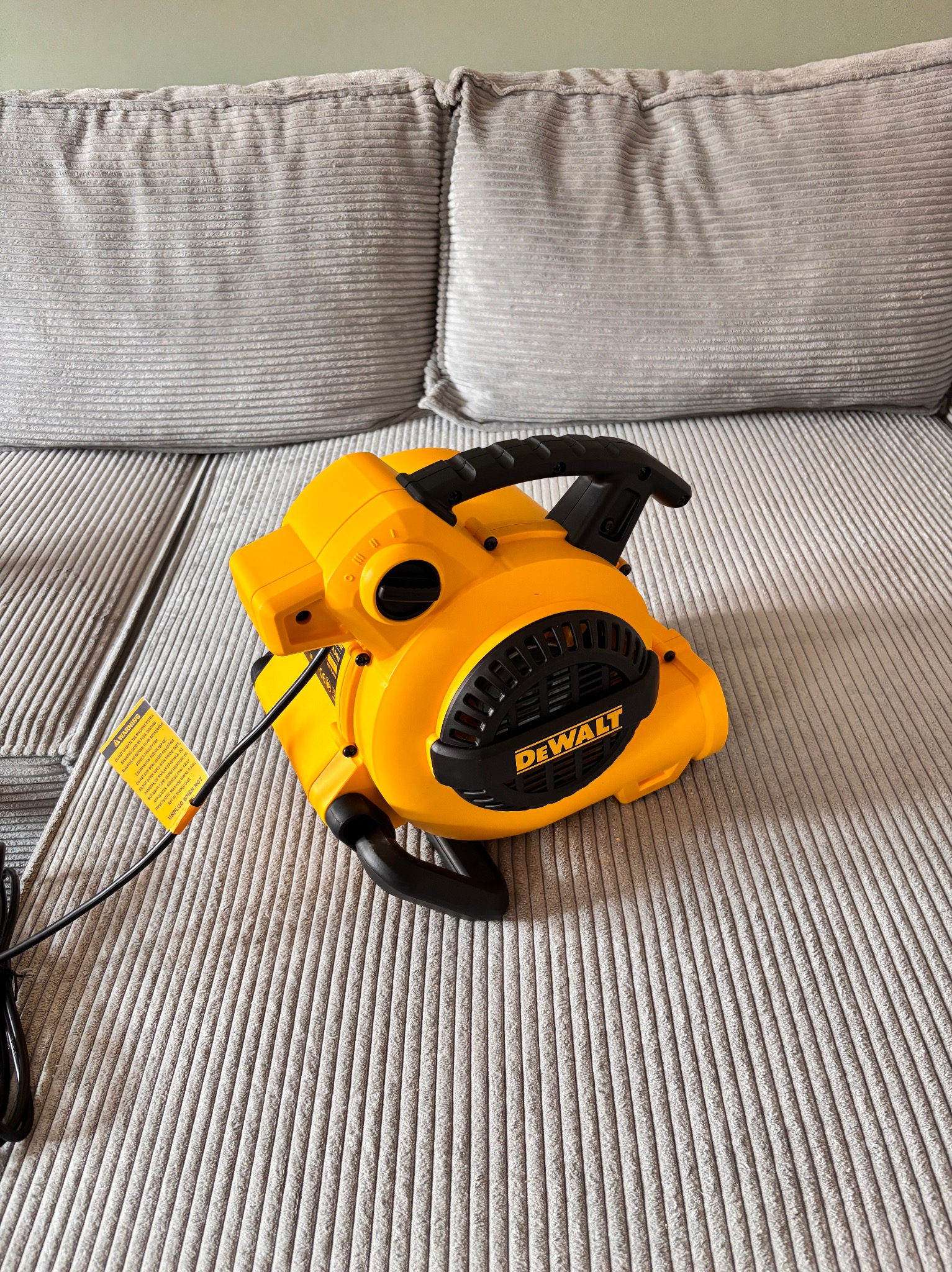 Żółty odkurzacz DeWalt na szarym, teksturowanym siedzisku sofy. Widoczne czarne elementy i etykieta ostrzegawcza. Sprzęt do czyszczenia tapicerki.