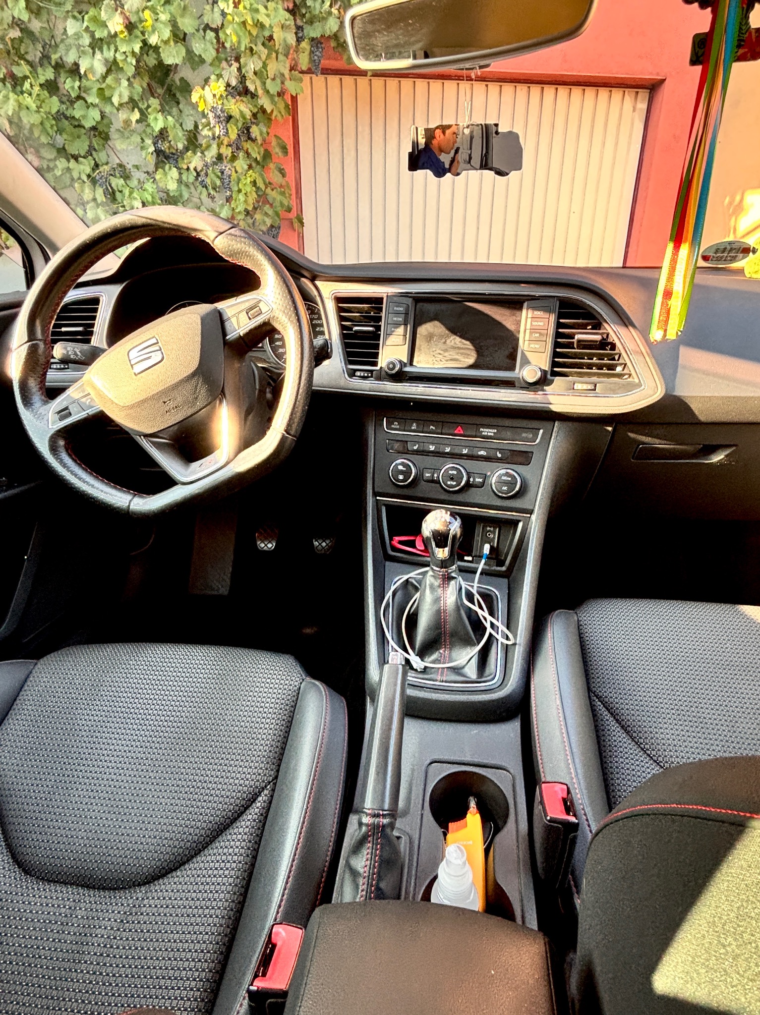 Wnętrze samochodu marki Seat z widoczną deską rozdzielczą, kierownicą i fotelami z czarną tapicerką i czerwonymi przeszyciami. W tle widoczne winogrona.