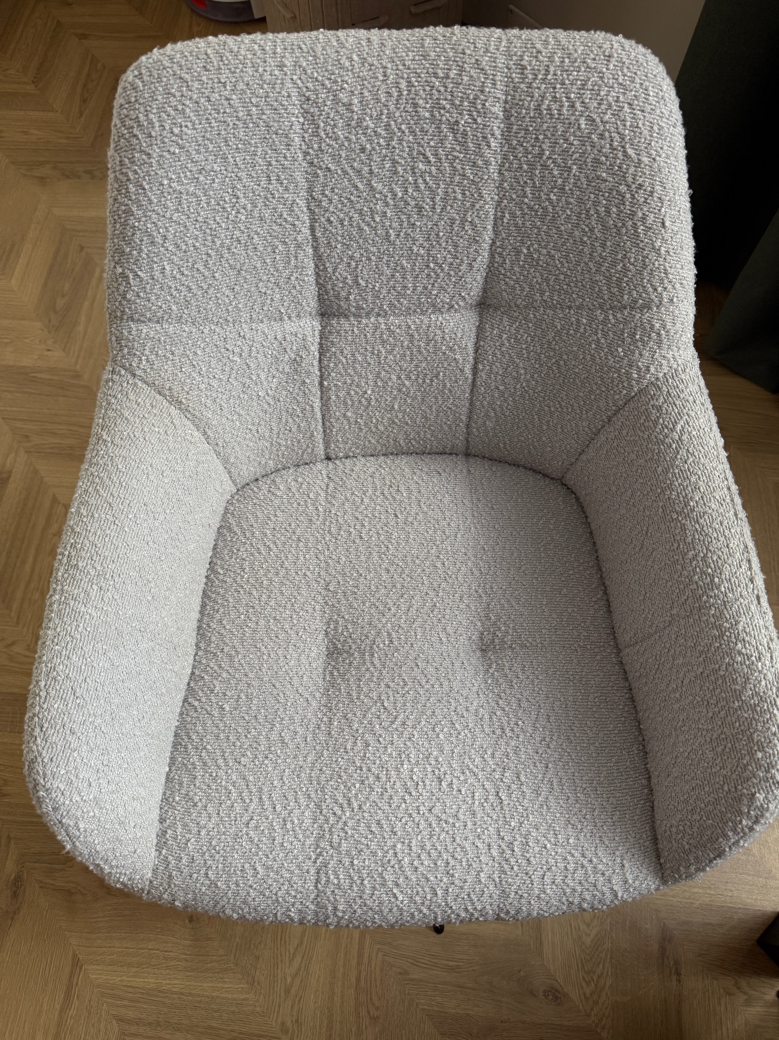 Kadr z góry na krzesło tapicerowane w jasnoszarym odcieniu, z widoczną fakturą materiału i subtelnymi przeszyciami, stojące na parkiecie ułożonym w jodełkę.