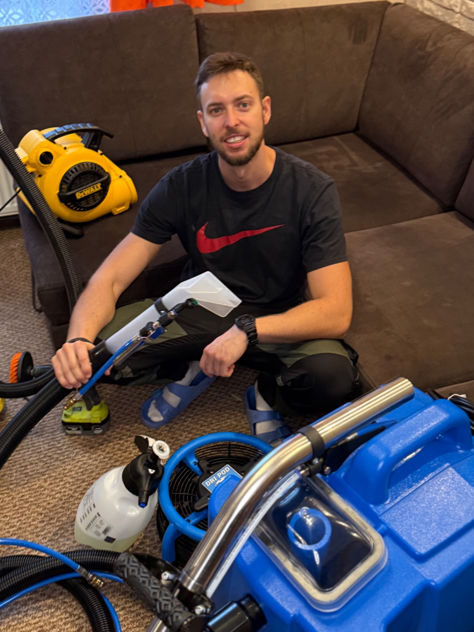 Uśmiechnięty mężczyzna siedzi na podłodze obok brązowej kanapy, otoczony sprzętem do czyszczenia tapicerki, w tym odkurzaczem i niebieskim urządzeniem Dri-Pod.