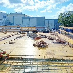 AST-BUD - Budowa płyty fundamentowej: widok z góry na zbrojenie siatką stalową, szalunki drewniane i stosy płyt betonowych na placu budowy z budynkiem w tle.