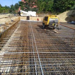 AST-BUD - Siatka zbrojeniowa na fundamencie, z żółtym kompresorem powietrza w tle i stertą białych płyt izolacyjnych po lewej stronie.