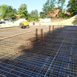 AST-BUD - Siatka zbrojeniowa fundamentu na czarnej folii, z wystającymi prętami pionowymi, żółty agregat prądotwórczy w tle, widok z góry.