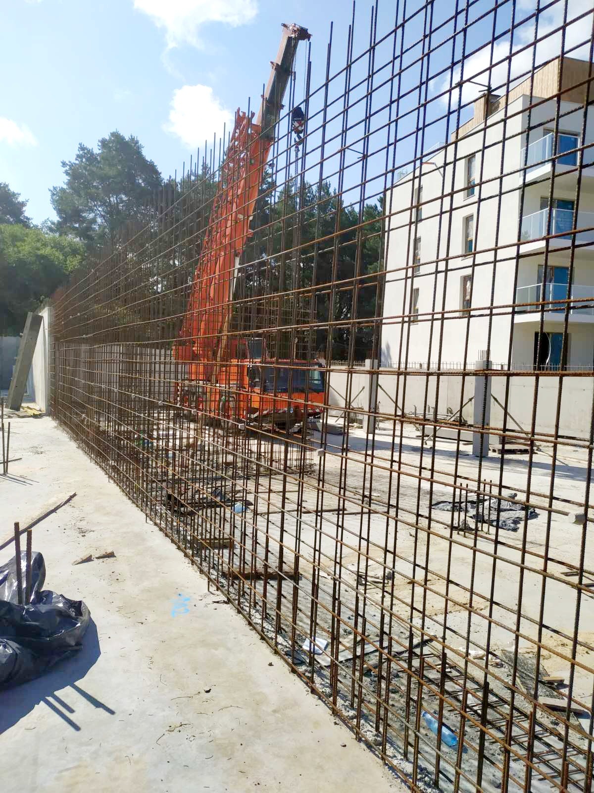 Zbrojenie pionowe ściany żelbetowej z widocznym dźwigiem budowlanym i fragmentem budynku w tle.