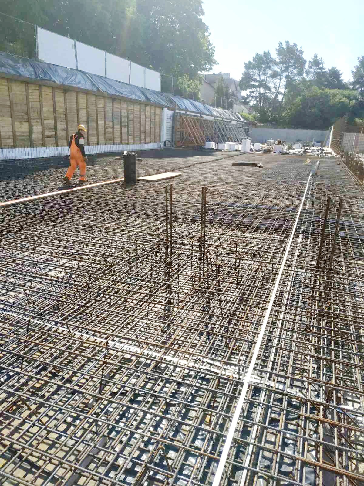 Siatka zbrojeniowa na fundamencie z wystającymi prętami, pracownik w pomarańczowym kombinezonie w tle, widoczne szalunki i przygotowania do wylania betonu.