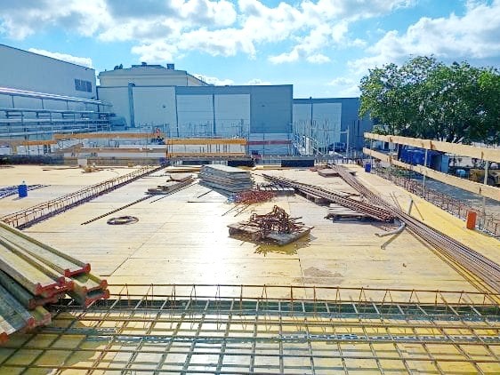 Budowa płyty fundamentowej: widok z góry na zbrojenie siatką stalową, szalunki drewniane i stosy płyt betonowych na placu budowy z budynkiem w tle.