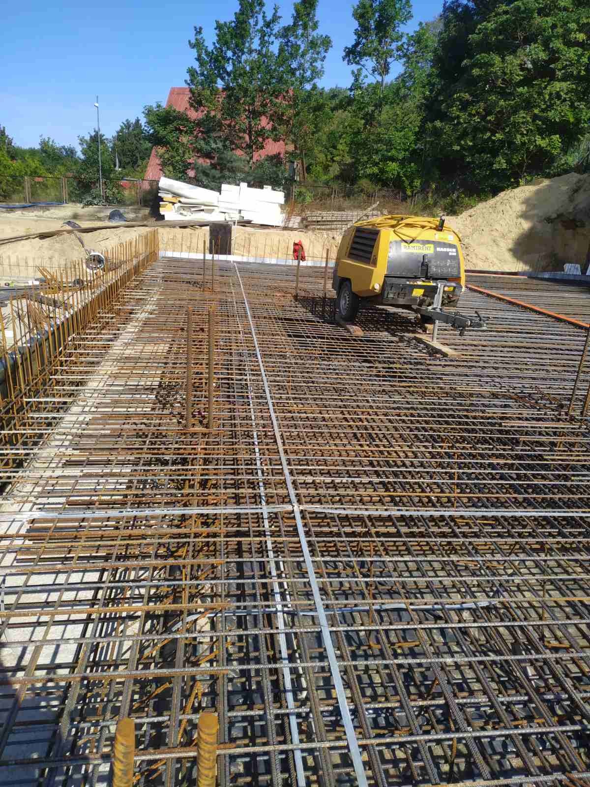 Siatka zbrojeniowa na fundamencie, z żółtym kompresorem powietrza w tle i stertą białych płyt izolacyjnych po lewej stronie.