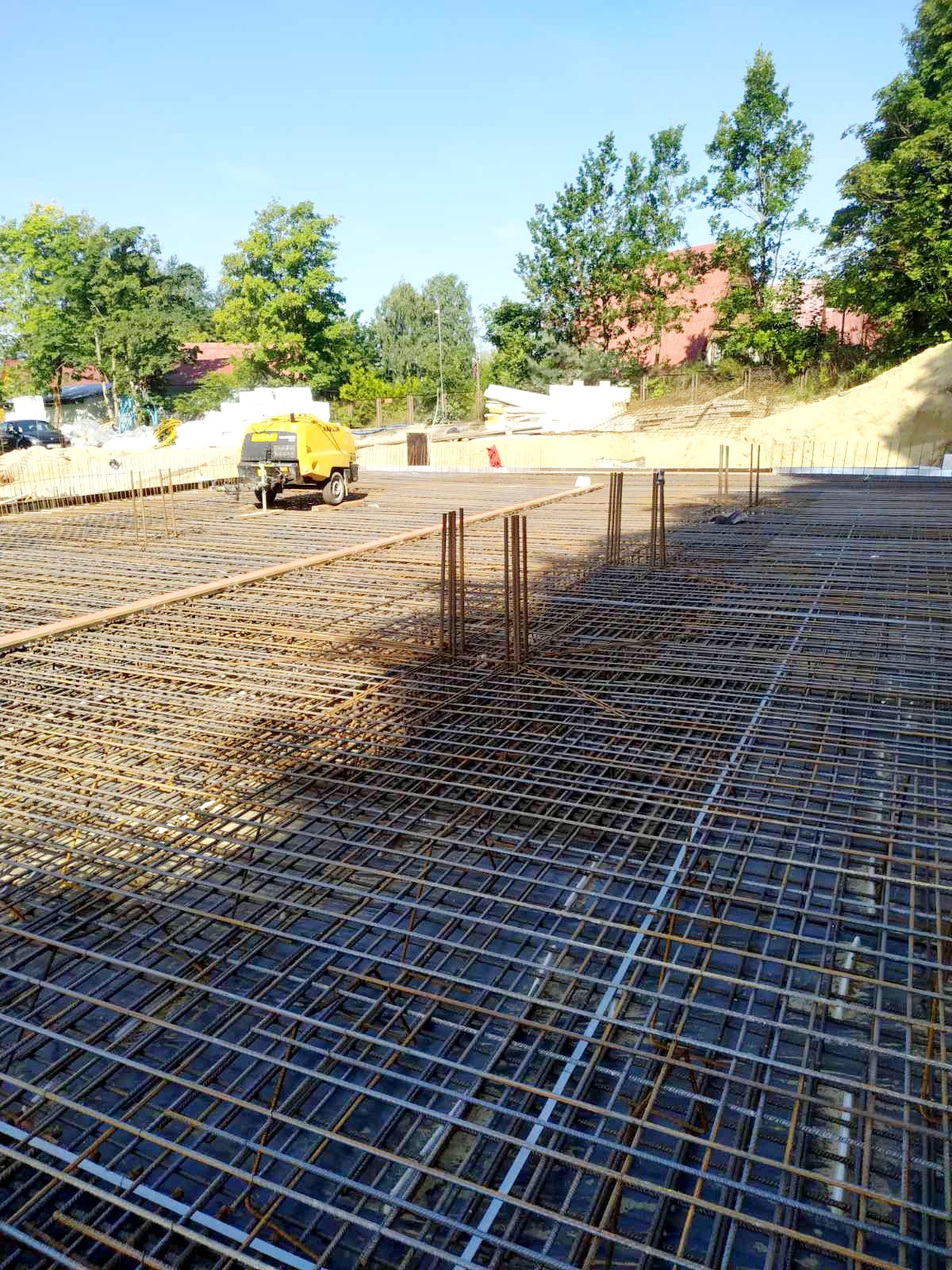 Siatka zbrojeniowa fundamentu na czarnej folii, z wystającymi prętami pionowymi, żółty agregat prądotwórczy w tle, widok z góry.