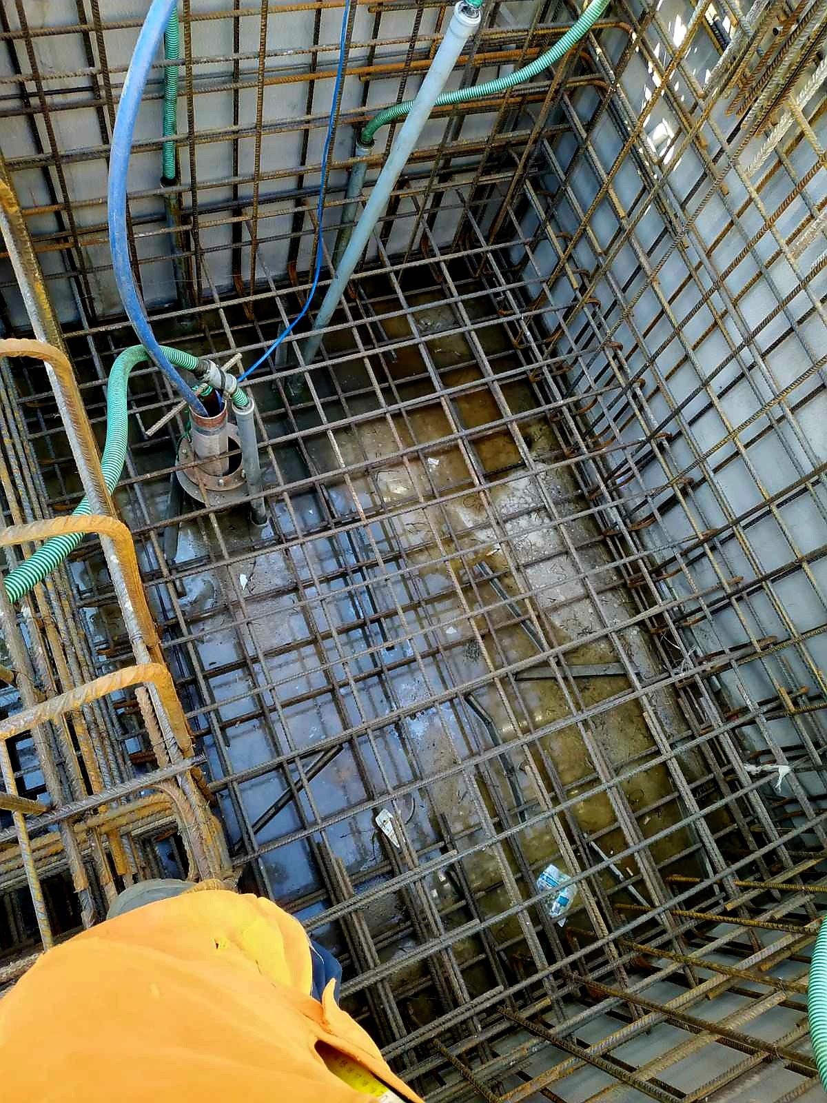 Siatka zbrojeniowa w wykopie fundamentowym, częściowo zalana wodą, z widocznymi przewodami i elementami instalacji. Widok z góry z fragmentem ubrania osoby.